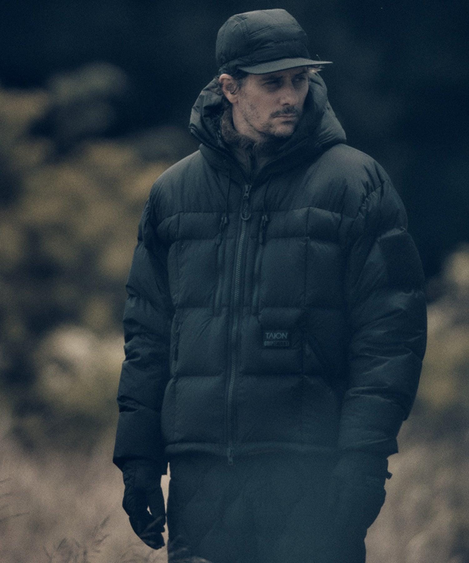 Hooded black GRIP SWANY × TAION jacket worn outdoors with cap.
