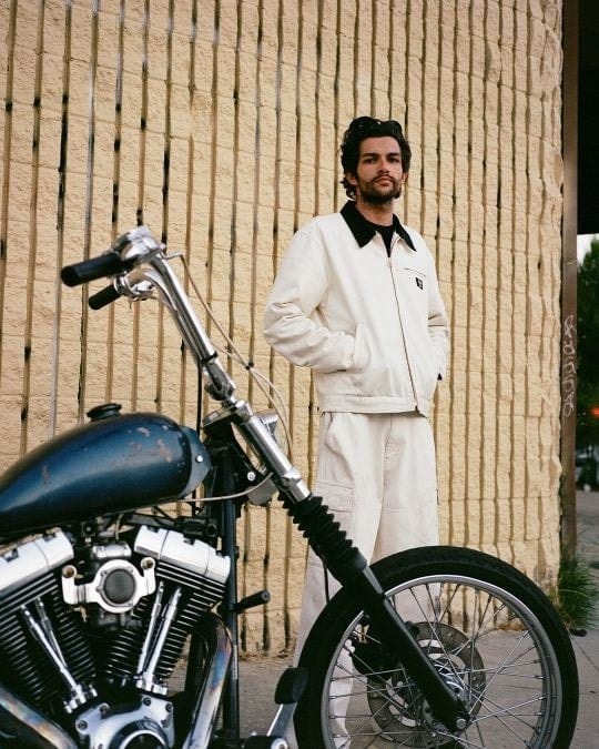 Model in a natural-coloured HUF and Dickies canvas jacket and trousers beside a motorcycle.